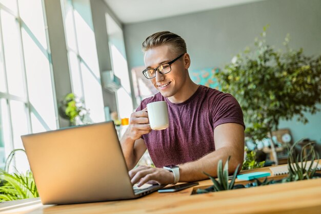 Foto giovane uomo d'affari allegro che beve caffè mentre usa il portatile in ufficio