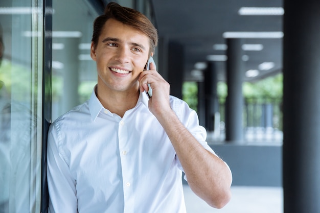 Cheerful young businessman holding laptop and talking on mobile phone near business center