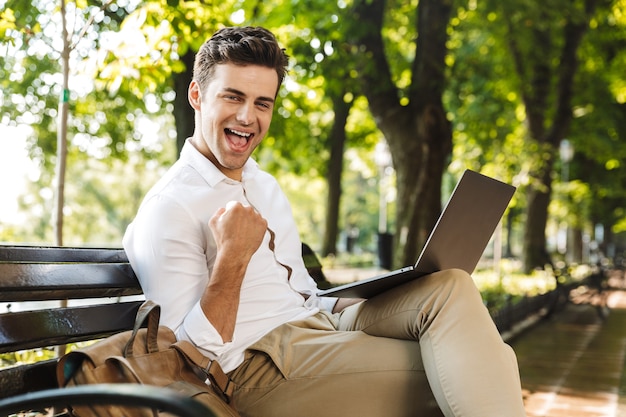 Cheerful young businessman sitting on a bench outdoors, working on laptop computer, celebrating success