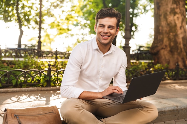 Cheerful young businessman sitting outdoors, working on laptop computer