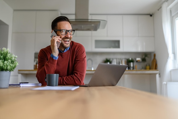 Cheerful young businessman talking over smart phone while working over laptop on desk in home office