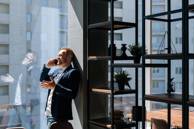 Cheerful young businessman wearing fashion suit is talking on mobile phone in modern office room near large window. Concept of office working.