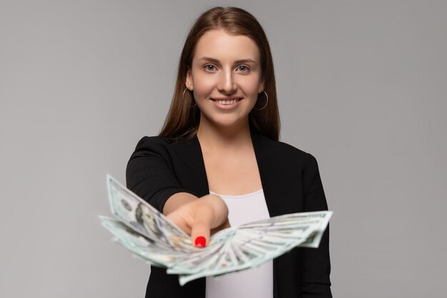 Foto giovane donna di affari allegra in vestito nero che dà le banconote del dollaro isolate su gray