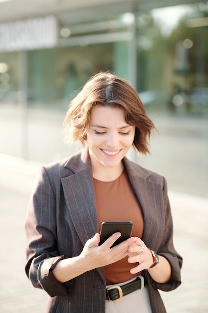 Cheerful young businesswoman in smart casualwear scrolling through contacts in smartphone outdoors while going to phone someone