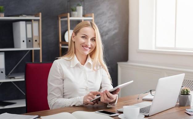 Cheerful young businesswoman using her touchpad with smile while sitting at her working place in office