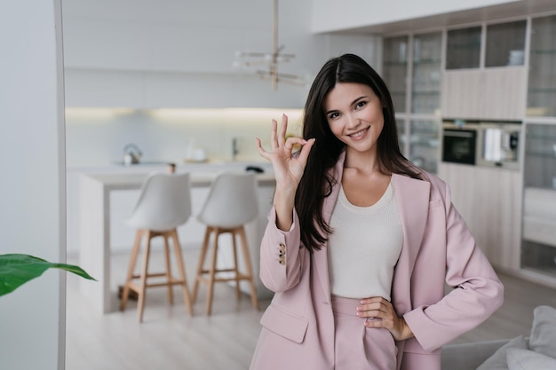 Cheerful young businesswoman with loose dark hair looking at camera toothy smiling gestures ok sign by hand standing over blurry living room Remote working Business people Happy student