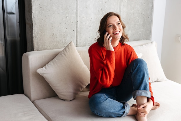 Cheerful young casually dressed woman sitting on a couch at home, talking on mobile phone