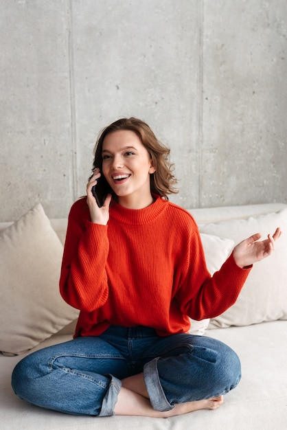 自宅のソファに座って、携帯電話で話している陽気な若いカジュアルな服装の女性