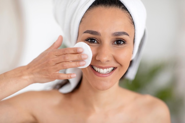 Photo cheerful young caucasian brunette woman in towel wipes face with cotton pad and lotion in light