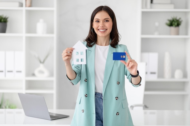 Веселая молодая кавказская деловая женщина в костюме держит кредитную карту и дом