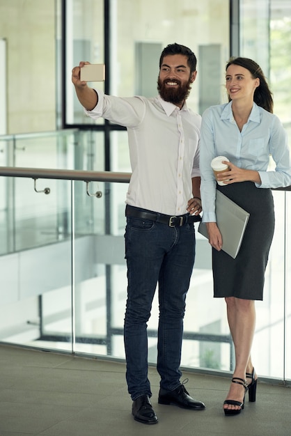 Cheerful young caucasian colleagues in smart casual outfits posing against railing in lobby and phot...