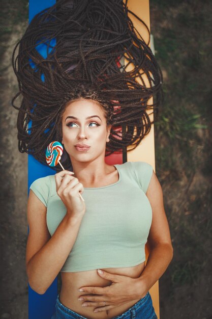 Cheerful young Caucasian woman with beautiful long afro braided hair is licking the lollipop, pouting her lips and looking away.