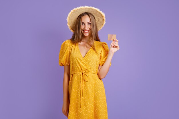 Cheerful young caucasian woman in yellow dress and straw hat holding bank card isolated on purple ba...