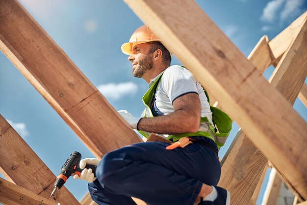 Cheerful young constructor sitting on the beam while preparing object for covering the roof