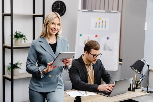 Cheerful young corporate lawyers in formal wear editing text of new contract using laptop sitting at table with laptop in modern office Woman sits next to a colleague and holds a tablet in her hands