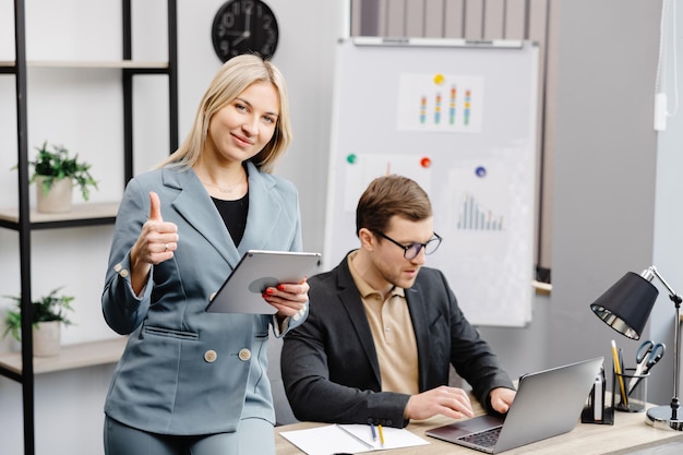 Cheerful young corporate lawyers in formal wear editing text of new contract using laptop sitting at table with laptop in modern office Woman sits next to a colleague and holds a tablet in her hands