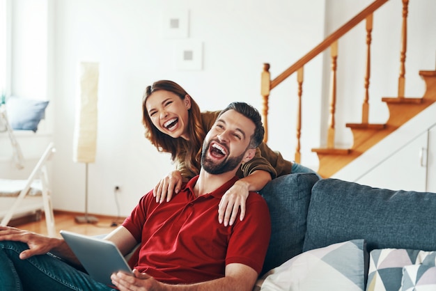 Веселая молодая пара в повседневной одежде сближается и смеется, отдыхая на диване в помещении