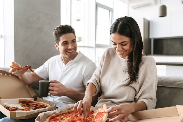 Allegro giovane coppia seduta su un divano a casa, mangiare la pizza