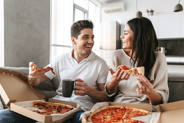 Веселая молодая пара, сидя на диване у себя дома, ест пиццу