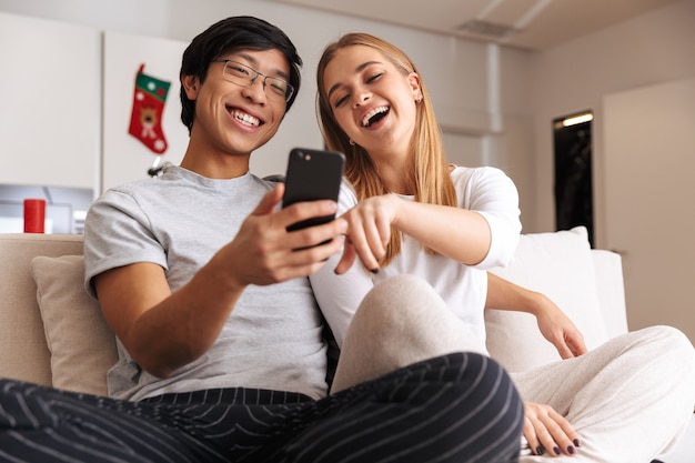 Cheerful young couple, sitting together on a couch at home, looking at mobile phone