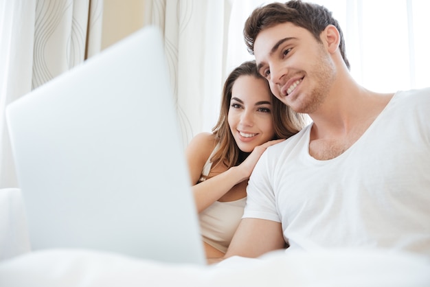 Cheerful young couple smiling and using laptop in bed