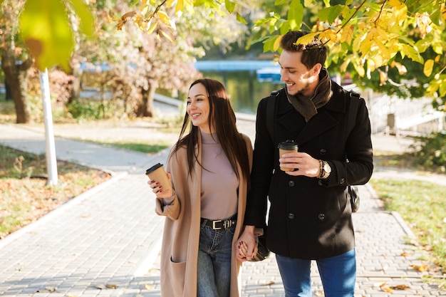 Cheerful young couple spending fun time at the park in autumn, walking holding hands, drinking takeaway coffee