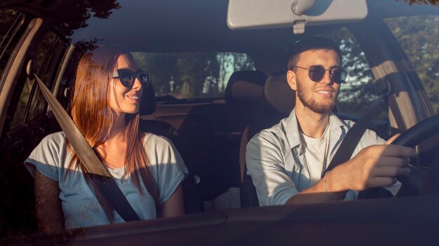 Cheerful young couple in sunglasses driving car through summer countryside in sunset