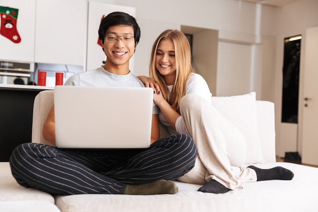 Веселая молодая пара, используя портативный компьютер, сидя на диване у себя дома