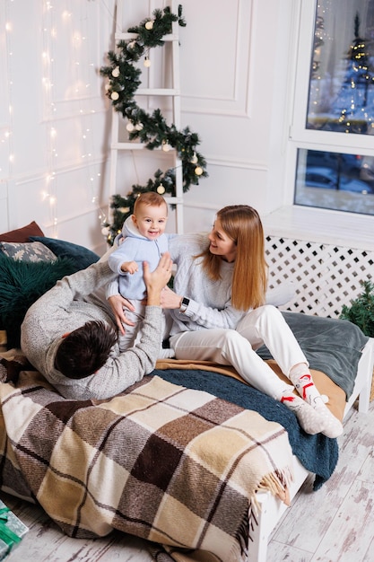 Una giovane coppia allegra con un figlio piccolo sta giocando sul letto vicino all'albero di natale interno di capodanno nella camera da letto albero di natale con i giocattoli atmosfera familiare festosa