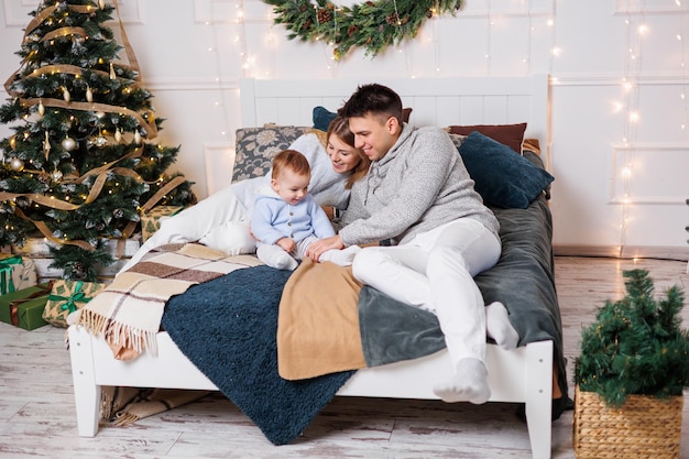 A cheerful young couple with a little son are playing on the bed near the Christmas tree New Year's interior in the bedroom Christmas tree with toys Festive family atmosphere