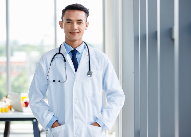 Medico maschio asiatico giovane e carino allegro in uniforme e con stetoscopio in piedi in clinica e posa con gesto positivo e sicuro di sé.