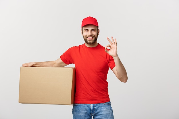 Cheerful young delivery man in red cap standing with parcel post box isolated over white