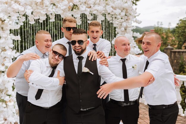 Cheerful young energetic witnesses of the groom next to the groom Friends congratulate the groom The groom in a vest and his friends in white shirts pose near the wedding arch Wedding in nature