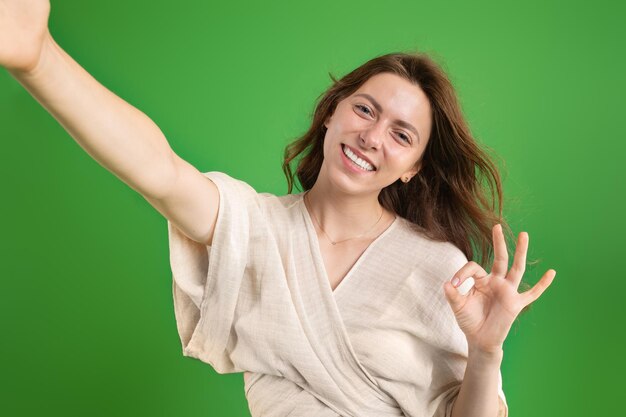 Cheerful young european lady take selfie ok sign isolated on green studio background