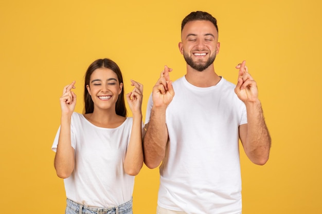 Cheerful young european man and woman in white tshirts with crossed fingers hoping or wishing