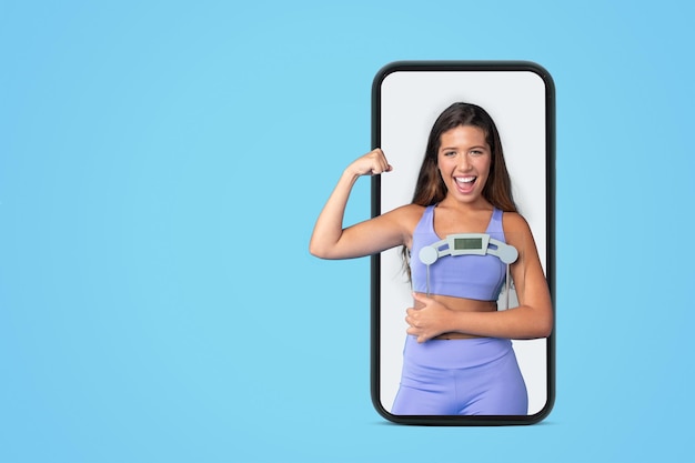 Cheerful young european slim lady in sportswear hold scales show muscles biceps on screen big phone