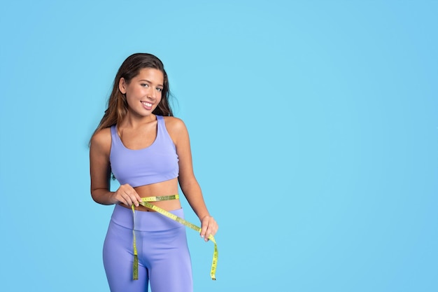 Cheerful young european slim woman athlete in sportswear measures waist with measuring tape