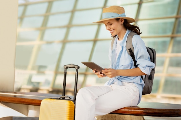 Cheerful young european woman in hat with suitcase and backpack typing on tablet