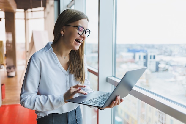 Cheerful young fairhaired business woman of European appearance in a white shirt and glasses holds a portable laptop in her hands stands near a large window in the office on the top floor