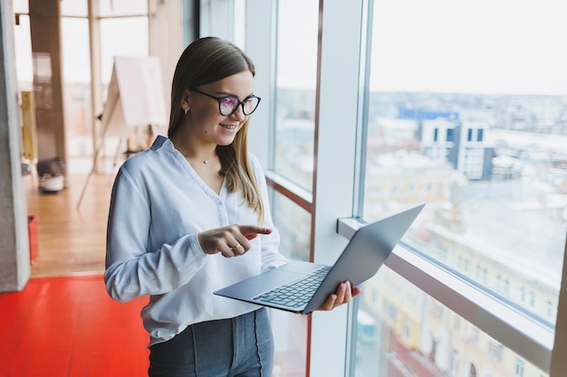 Cheerful young fairhaired business woman of European appearance in a white shirt and glasses holds a portable laptop in her hands stands near a large window in the office on the top floor
