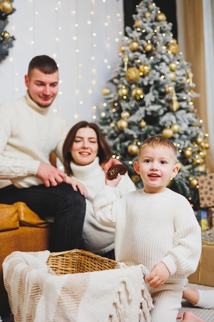 어린 아이를 데리고 있는 명랑한 젊은 가족은 집에 있는 크리스마스 트리 근처에 쉬고 있습니다. 집에 크리스마스 장식을 한 가족 커플 크리스마스 새해를 축하할 시간