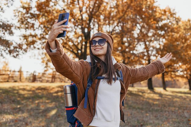 秋の森でのハイキング中に携帯電話のセルフポートレートを撮る陽気な若い女性バックパッカー