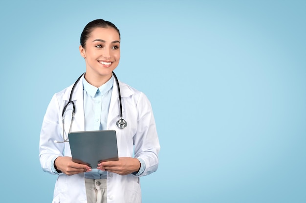 Cheerful young female doctor in white coat holding digital tablet looking at copy space over blue