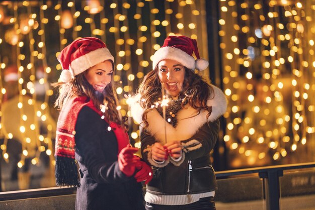 Cheerful young female friends are having fun in city street at night. They are holding sparklers and playing with it in holiday winter night.