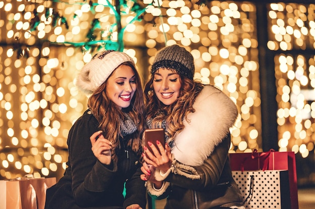 Photo cheerful young female friends are having fun in city street at night. they are surfing on social network at smartphone and talking something funny.