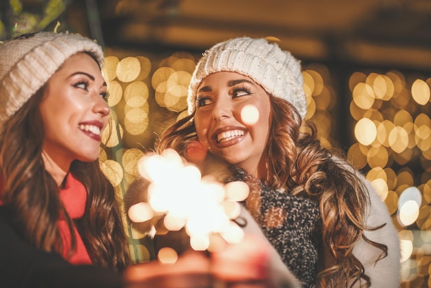 Cheerful young female friends are holding sparklers and having fun at holiday winter night in the city street.