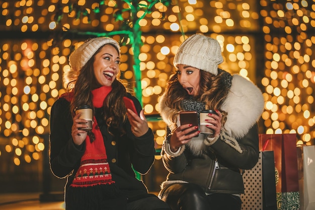 Cheerful young female friends are surfing on a social network at smartphone and having fun at holiday winter night in the city street.