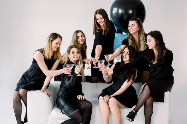 Cheerful young female friends in elegant black dresses with martini drinks enjoying birthday party or New Year celebration on white sofa at light room