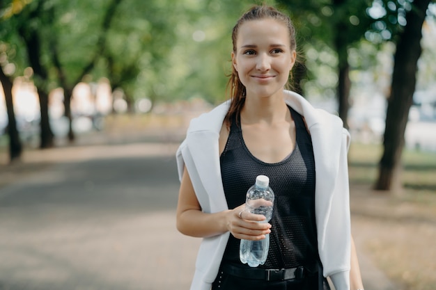 Веселая молодая бегунья держит позы из бутылки с водой на открытом воздухе в активной одежде, отдыхает после тренировки, прогулки в парке, освежаются после бега, чувствует жажду, поддерживает форму с обычным спортом