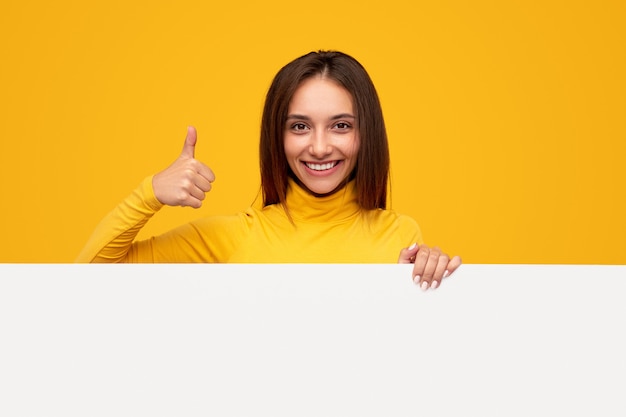 Cheerful young female smiling for camera and approving blank banner with thumb up gesture during advertisement campaign against yellow background